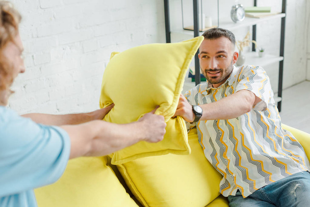 selective focus of cheerful homosexual men pillow fighting in living room  - Photo, Image