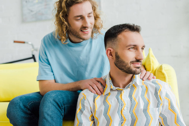Ευτυχισμένοι ομοφυλόφιλοι άντρες κοιτάζουν αλλού ενώ κάθονται στο σαλόνι.   - Φωτογραφία, εικόνα