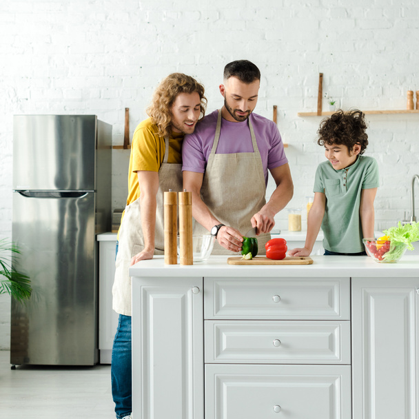 キッチンで調理するハンサムな同性愛者の両親の近くの幸せな混合レースの子供  - 写真・画像