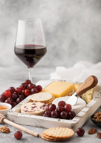 Glass of red wine with selection of various cheese in wooden box and grapes on light table background. Blue Stilton, Red Leicester and Brie Cheese with Cheddar and nuts with honey.  - Foto, afbeelding