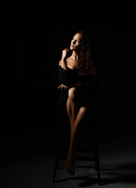 Sensual woman with long curly brunette hair in black off-shoulder dress is sitting on a high chair in darkness - Foto, imagen