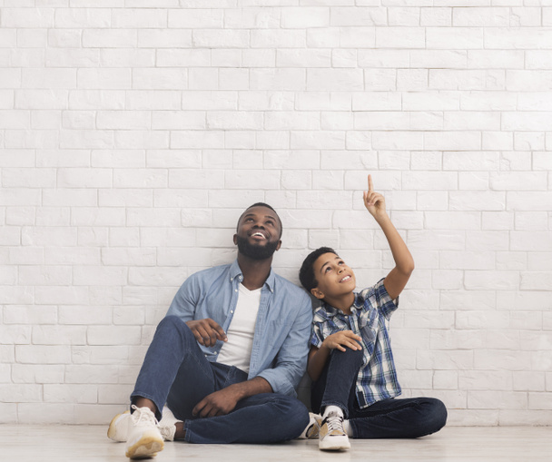 Father and son sitting on floor near white brick wall, pointing and looking upwards - Photo, image