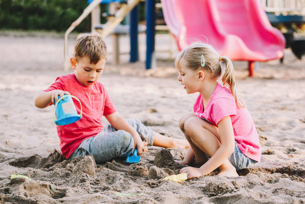 Two Caucasian children sitting in sandbox playing with beach toys. Little girl and boy friends having fun together on playground. Summer outdoor activity for kids. Leisure time lifestyle childhood. - Foto, Bild