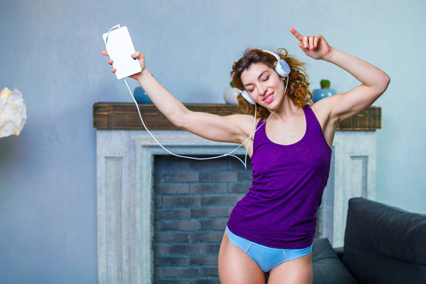 Cheerful young woman in a purple top and blue pants with headphones on is dancing to the music at home - Photo, image