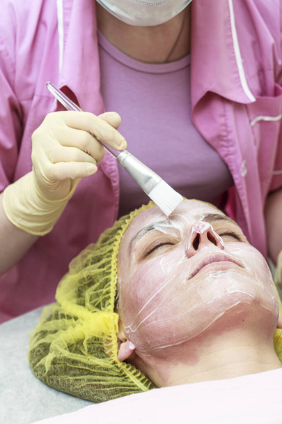 Kosmetikerin trägt nach chemischem Peeling eine beruhigende, weiße Maske auf die gerötete Haut des Patienten auf. - Foto, Bild