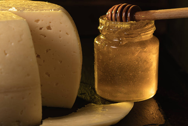 La cabeza de queso yace en un plato de pizarra que yace sobre una superficie oxidada. Muy cerca se encuentra un frasco manchado de miel en el que la barra de agitación de miel. Un trozo de queso cortado por una marca. oscuro
 - Foto, Imagen