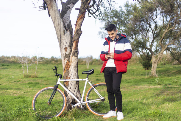 Handsome hipster young man enjoying a bicycle ride and using a smartphone while walking on park - Φωτογραφία, εικόνα
