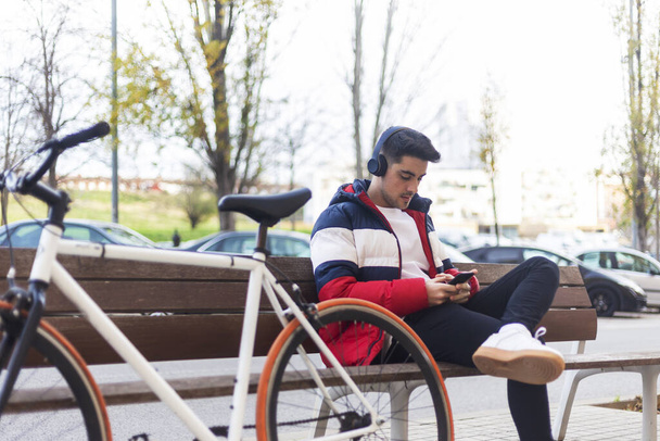 Young man sitting on a bench in the street while using mobile phone and listening music by headphones - 写真・画像