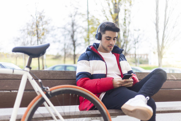Young man sitting on a bench in the street while using mobile phone and listening music by headphones - Photo, Image