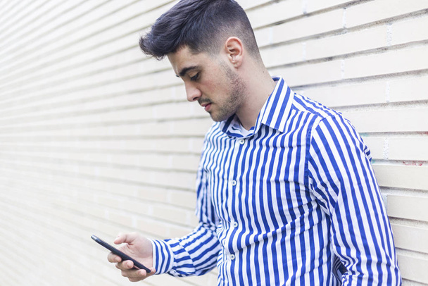 Young happy man leaning on house wall while using his smartphone - Foto, Imagen