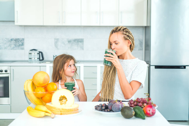 Happy woman mother and little girl daughter drinks green smoothie in kitchen. Mother and daughter with glass of natural detox smoothie in kitchen. Food supplements. - Foto, imagen