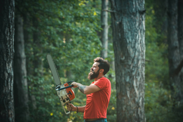Lumberjack in the woods with chainsaw axe. Firewood as a renewable energy source. Stylish young man posing like lumberjack. Lumberjack holding the chainsaw. - Photo, image
