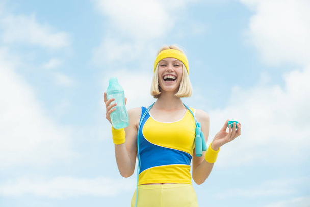 Beautiful blonde woman girl doing sport activities outdoor at fresh air. Sport is good for health. Cheerful smiling sporty girl with bottle of water at blue cloudy sky background. - Photo, Image