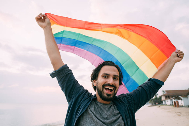 Chico alegre con una bandera de arco iris en la playa
.  - Foto, Imagen