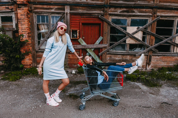 A long haired woman wearing a powder blue dress, pink woollen hat is chilling with her female friend, who is sitting on a shopping trolley, wearing a black leather jacket and blue jeans, in a derelict - Photo, Image