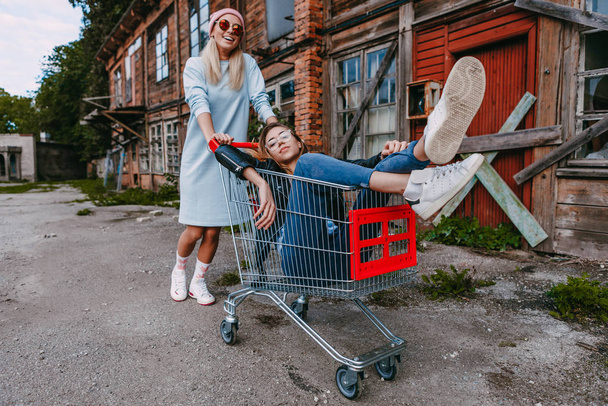 A long haired woman wearing a powder blue dress, pink woollen hat is chilling with her female friend, who is sitting on a shopping trolley, wearing a black leather jacket and blue jeans, in a derelict - Foto, Imagem