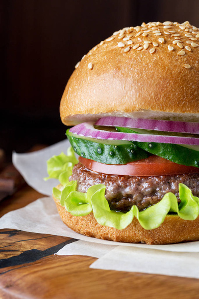 Burger on wood board at dark background. Sesame bun with beef, lettuce, tomato, cucumber and onion - Fotografie, Obrázek