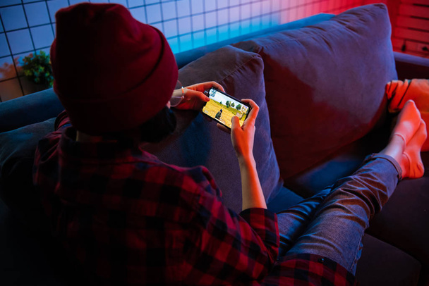 Moskva, Rusko - prosinec 2019: pohled zblízka na ženské ruce hrající online hru na chytrém telefonu ležícím doma na gauči - Fotografie, Obrázek