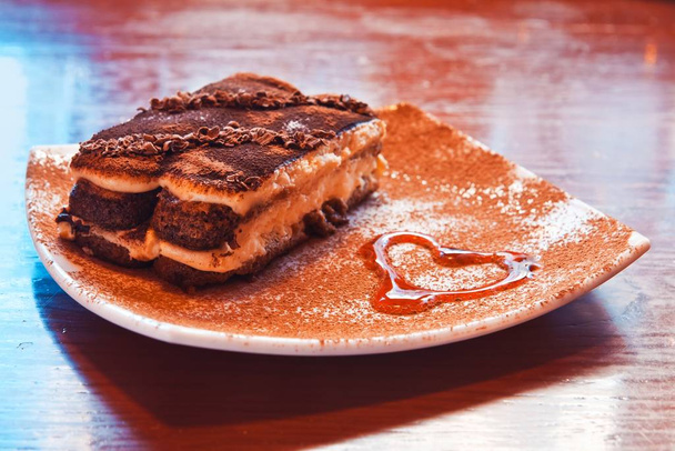 tiramisu dessert taart bestrooid met cacaopoeder en geserveerd op een bord met siroop geschilderd hart symbool in cafe, zoet heerlijk eten in romantische sfeer - Foto, afbeelding