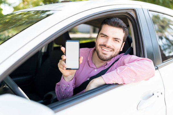 Hombre adulto feliz mostrando teléfono inteligente con pantalla en blanco mientras está sentado en un coche nuevo
 - Foto, Imagen