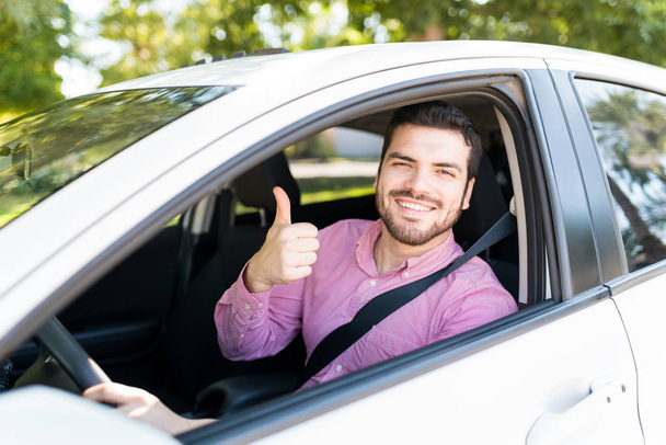 Portrait of smiling handsome man showing thumbs up while driving new car - 写真・画像