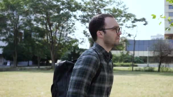 A handsome young man in glasses and with backpack walking along the park. - Filmati, video