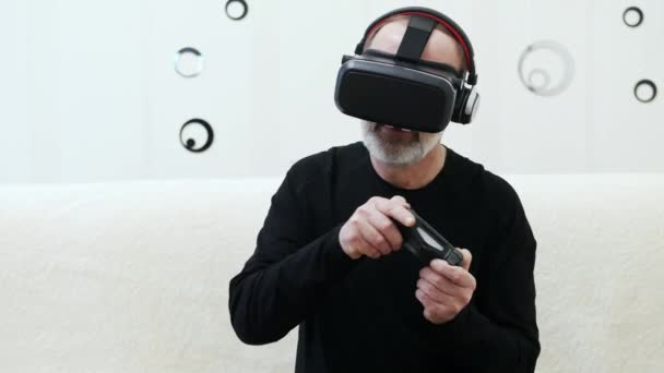 Hombre de mediana edad con barba gris juega en un videojuego de realidad virtual con auriculares inalámbricos, gamepad y auriculares VR
 - Metraje, vídeo