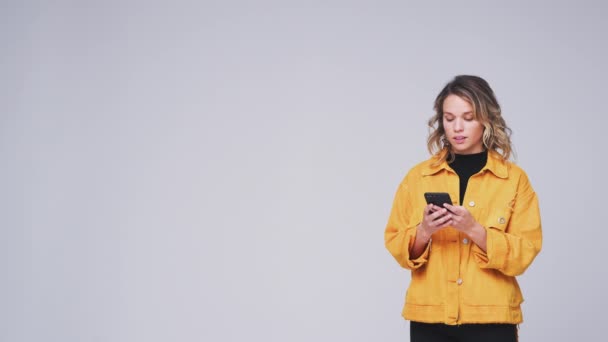 Young woman sending text message on mobile phone looks up against white studio background - shot in slow motion - Felvétel, videó