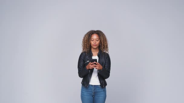 Young woman wearing leather jacket sending text message on mobile phone against white studio background - shot in slow motion - Filmati, video