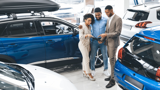 Couple Visiting Car Dealership Talking With Salesman In Auto Showroom - Foto, imagen