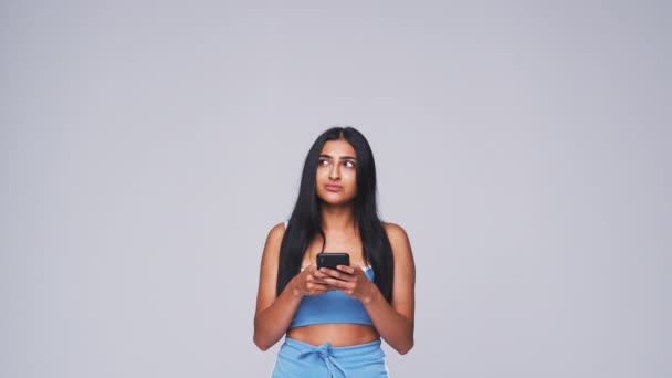 Young woman sending text message on mobile phone looks up against white studio background - shot in slow motion - Footage, Video