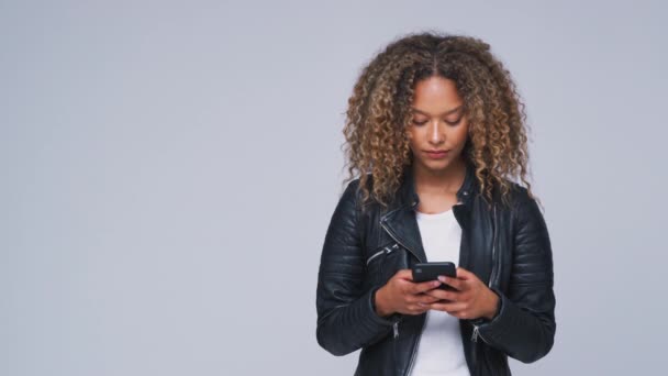 Young woman wearing leather jacket sending text message on mobile phone looks up against white studio background - shot in slow motion - Séquence, vidéo