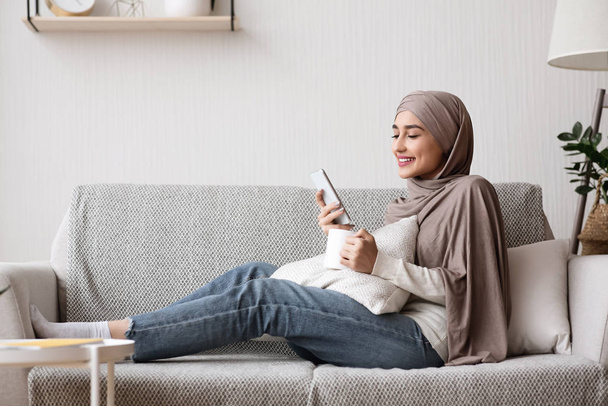 Young Arab Housewife Relaxing At Home With Smartphone And Coffee - 写真・画像