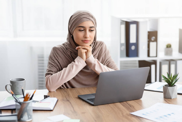 Nachdenkliche muslimische Geschäftsfrau sitzt am Arbeitsplatz und blickt auf den Laptop-Bildschirm - Foto, Bild