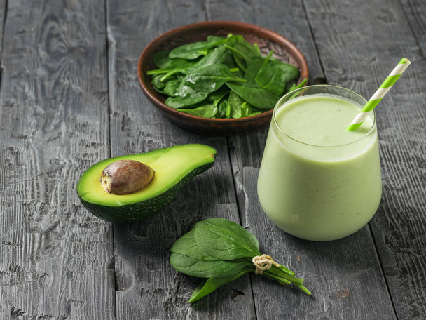 A bowl of spinach leaves, half an avocado, and a glass of smoothies on a black wooden table. Fitness product. - 写真・画像