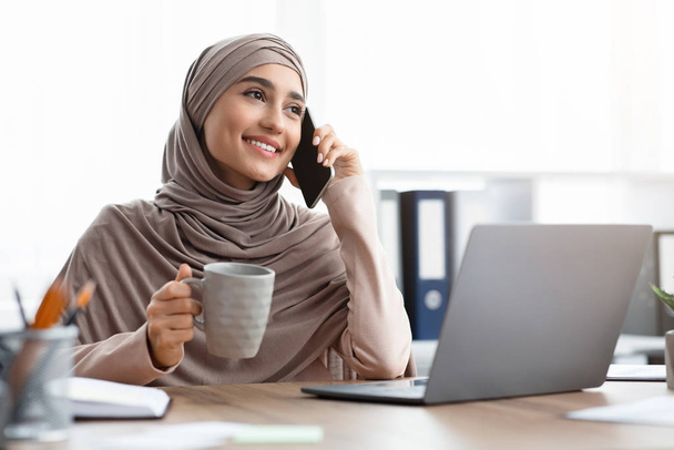 Heureuse femme d'affaires arabe parlant sur son téléphone portable au bureau et buvant du café
 - Photo, image