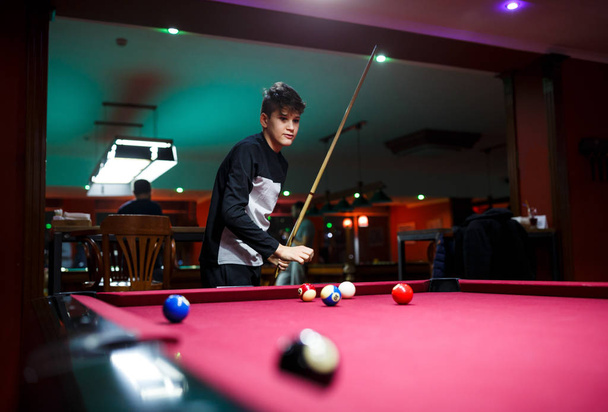 Boy plays billiard or pool in club. Young Kid learns to play snooker. Boy with billiard cue strikes the ball on table. Active Leisure, sport, hobby concept. - Photo, image
