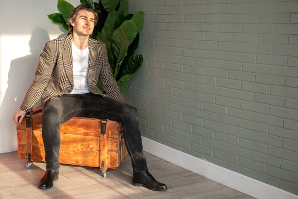 Tall handsome man sitting on the wooden box next to green plant and grey wall - Photo, image