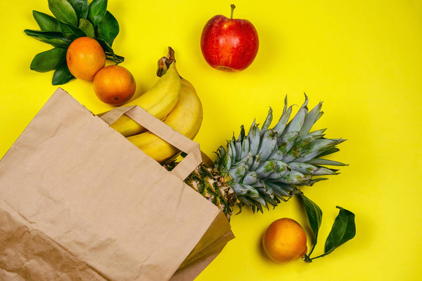top view flat lay with paper eco bag full of tropical fruits bananas,tangerines, apples and pineapple on a vibrant yellow background - Foto, Imagem