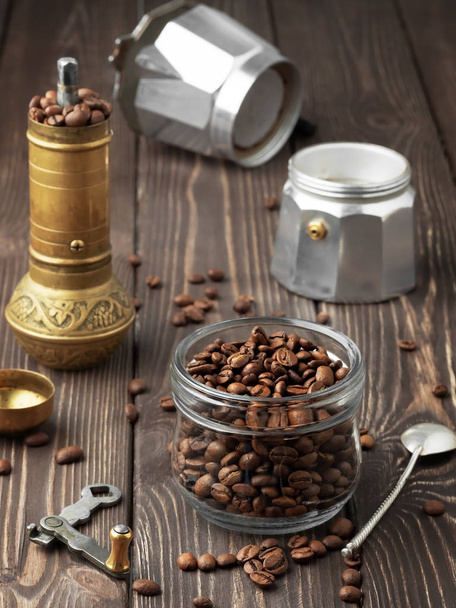 Pieczone ziarna kawy w szklanym słoiku i rozrzucone ziarna kawy na drewnianym brązowym stole. Młyn ręczny i dzbanek do kawy (la moka) w tle. Close Up, koncepcja tradycyjnego przygotowania kawy. - Zdjęcie, obraz