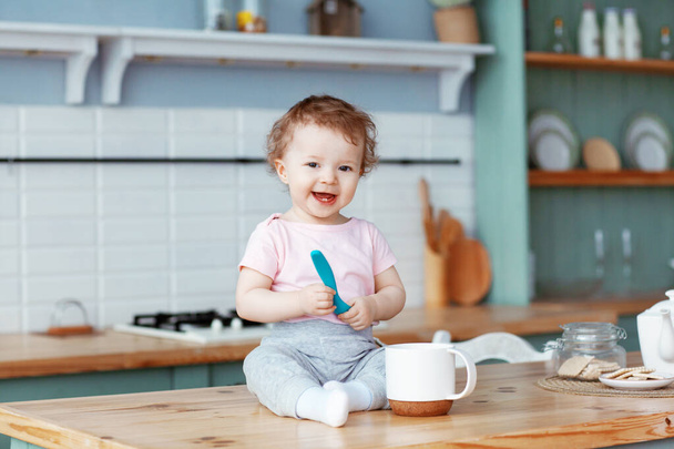 テーブルの上のキッチンに座っている幸せな赤ちゃん彼の手の中に食べ物のためのプラスチック製のスプーンを保持し、幅広く笑顔 - 写真・画像