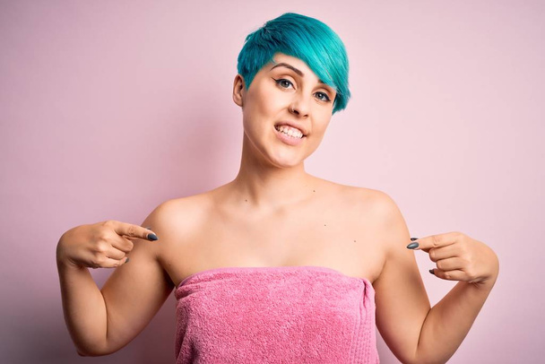Jovem mulher com cabelo azul moda vestindo toalha após o chuveiro sobre fundo rosa olhando confiante com sorriso no rosto, apontando-se com os dedos orgulhosos e felizes
. - Foto, Imagem