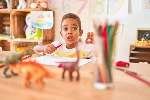 Hermoso niño afroamericano sentado dibujo usando papel y lápices en el escritorio en el jardín de infantes
 - Foto, imagen