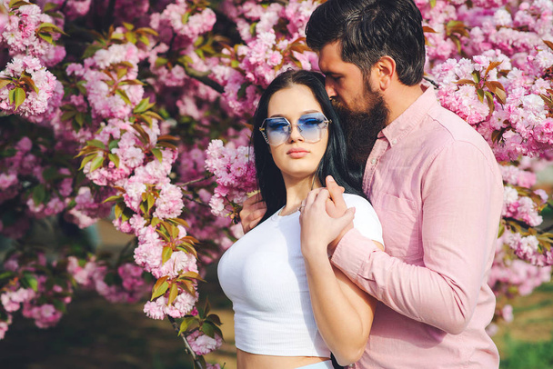 Loving bearded man and woman in a spring park. Spring pink sakura blossom. Pretty woman and her handsome boyfriend hugging outdoors. - 写真・画像