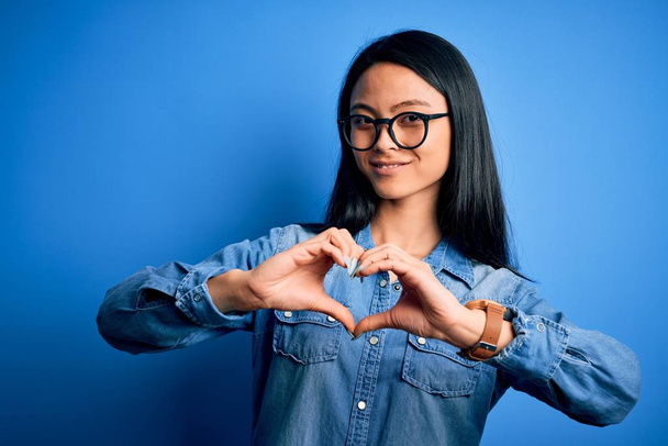 Молодая красивая китаянка в джинсовой рубашке на изолированном голубом фоне улыбается в любви, показывая символ сердца и форму руками. Романтическая концепция
. - Фото, изображение