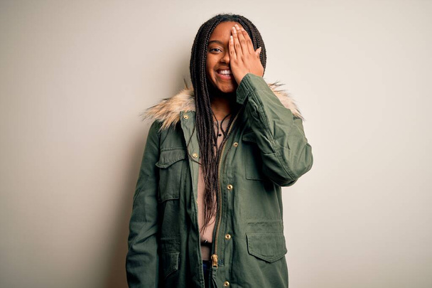 Giovane donna afroamericana che indossa il parka invernale su sfondo isolato che copre un occhio con mano, sorriso fiducioso sul viso ed emozione sorpresa
. - Foto, immagini