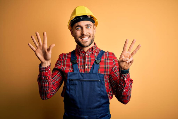 Jonge bouwvakker draagt bouwuniform en veiligheidshelm over gele geïsoleerde achtergrond die met vingers nummer acht naar boven wijst en zelfverzekerd en gelukkig glimlacht. - Foto, afbeelding