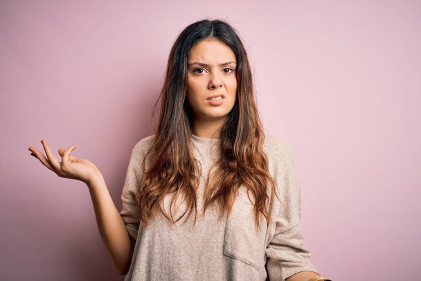 Young beautiful brunette woman wearing casual sweater standing over pink background clueless and confused with open arms, no idea concept. - Photo, Image