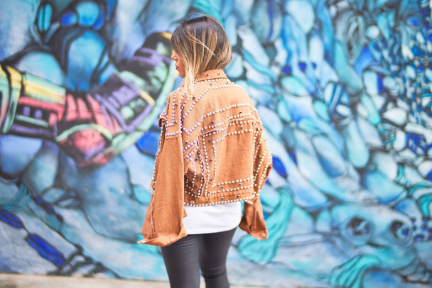 Молода красива дівчина в коричневій куртці стоїть позаду барвистої стіни на міській вулиці
 - Фото, зображення