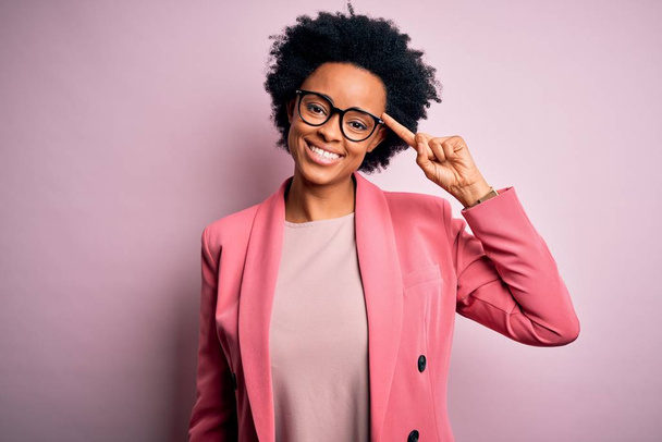 Fiatal gyönyörű afro-amerikai üzletasszony göndör haj visel rózsaszín kabát mosolygó mutató fej egy ujj, nagy ötlet vagy gondolat, jó memória - Fotó, kép